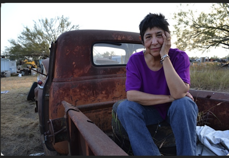 A still from Antonia: A Chicana Story. showing Antonia Castañeda wearing a purple shirt, sitting in the back of a vintage red pick up truck. She is facing toward the camera, smiling with her head resting on her hand.